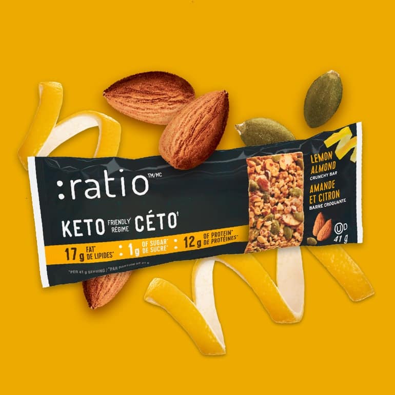 A Lemon Almond Ratio bar surrounded by almonds, lemon peels, and pumpkin seeds on a bright yellow background