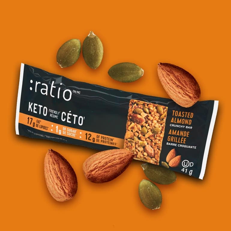 A Toasted Almond Ratio bar surrounded by almonds and pumpkin seeds on a bright orange background